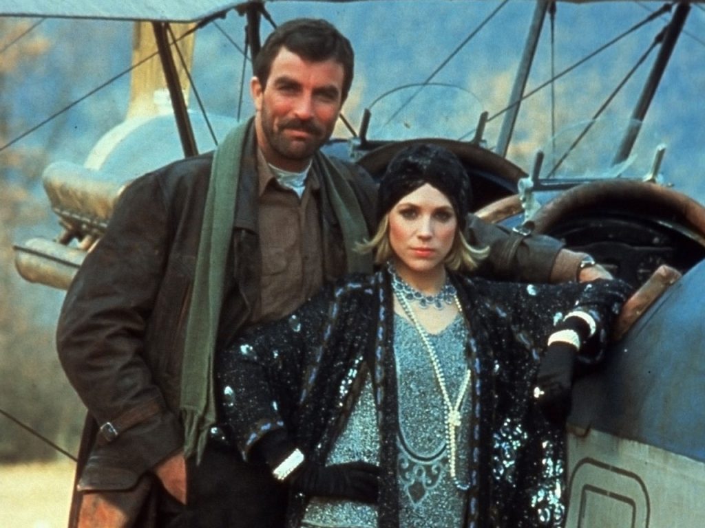 Tom Selleck and Bess Armstrong standing next to a bi-plane in High Road to China